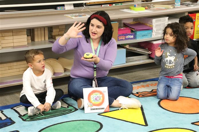  Image of pre-k teacher holding up her hand and teaching to a group of students